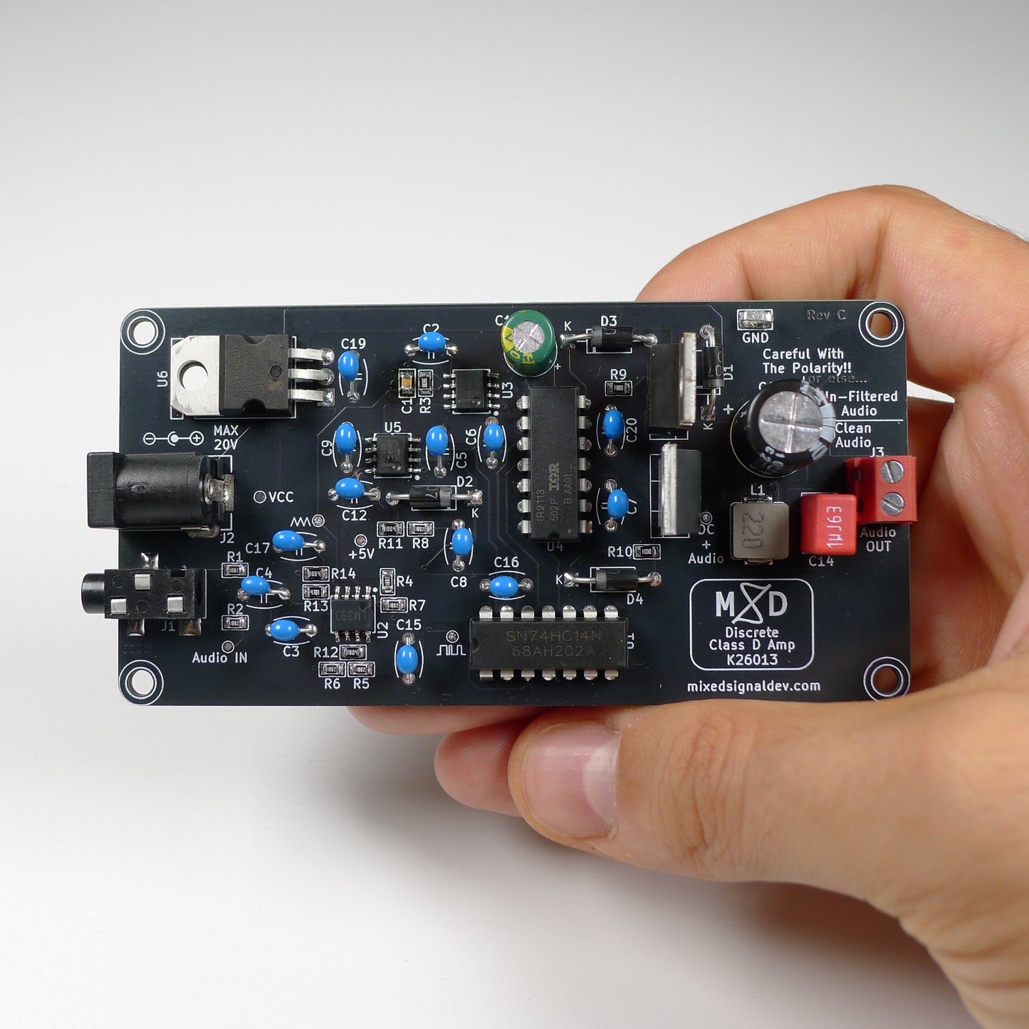 A hand holding a black electronic circuit board with various electronic components, including blue potentiometers, a green LED, and a white speaker, against a light gray background.