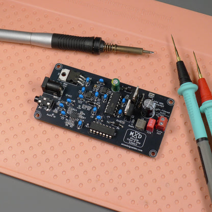 An electronic circuit board with various electronic components, shown on a peach-colored textured mat alongside a soldering iron and multimeter probes.